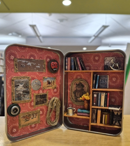 Miniature bookshelf built in a tin featuring red wallpaper, small books, and tiny art.